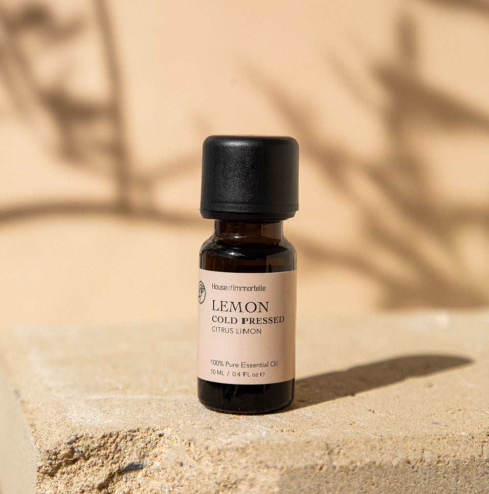 Clear glass bottle of lemon essential oil with a bright yellow label, surrounded by fresh lemon slices and green leaves, showcasing its refreshing, citrusy fragrance